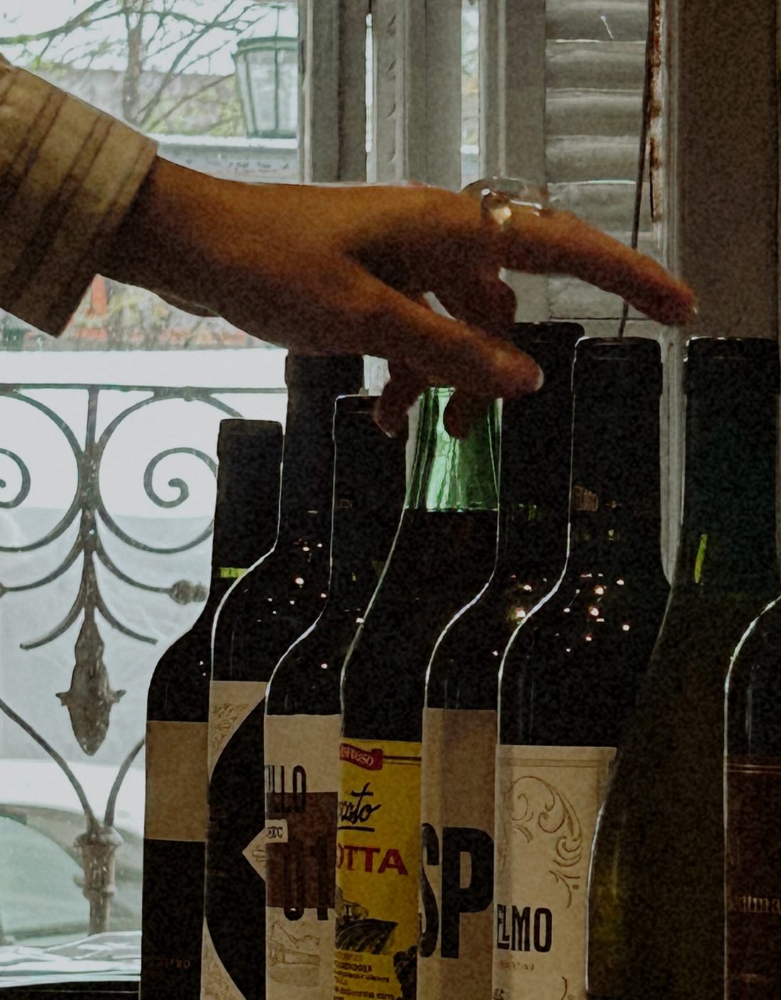 Una mano elige un vino entre las botellas, junto a la ventana con reja que da a la Plaza Dorrego